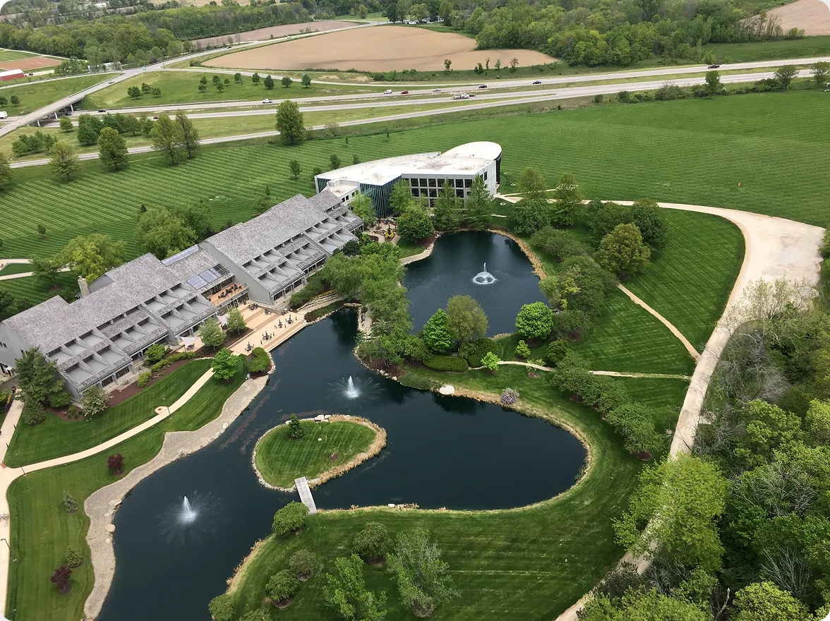 Aerial view of the ScottsMiracle-Gro campus showing a office building next to a curved pond with fountains, surrounded by green lawns and trees. Text: "We're all about green grass... And 'weed-free' is a very strong word here."