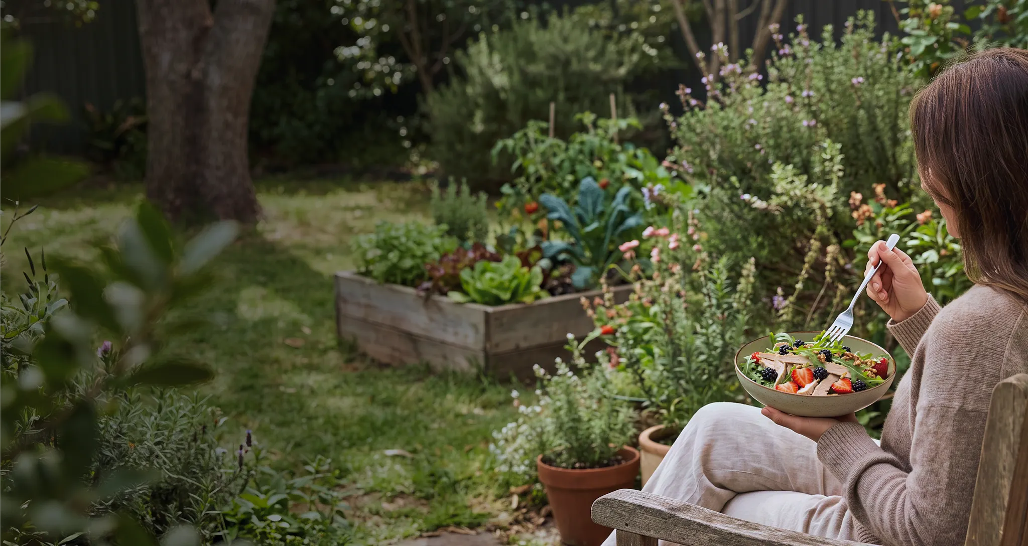 An overhead, warm-lit photograph of a person sitting outdoors on a wooden chair next to a garden bed, eating a salad from a bowl with a fork. Overlaid text on the upper left reads, "3 High-protein salads you'll absolutely crave," followed by a subhead encouraging gathering fresh garden ingredients, and a byline reading "By: Samantha Cassetty, MS RD".