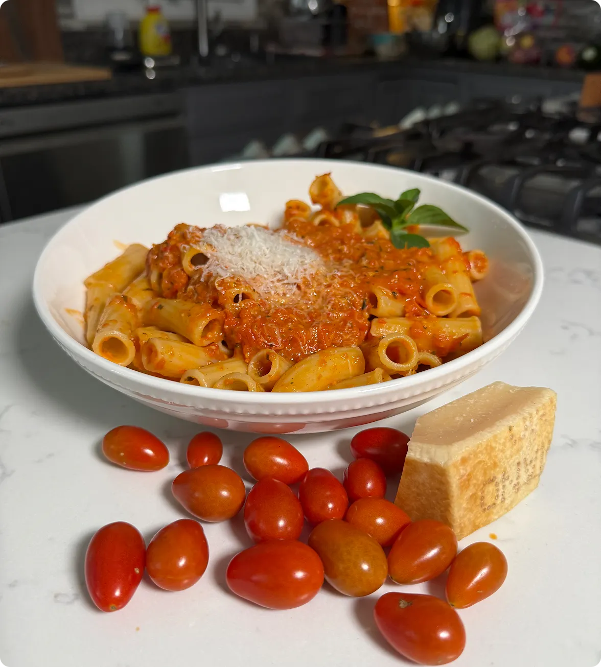 Bowl of pasta tossed in tomato sauce topped with basil, surrounded by fresh tomatoes and parmesan cheese.