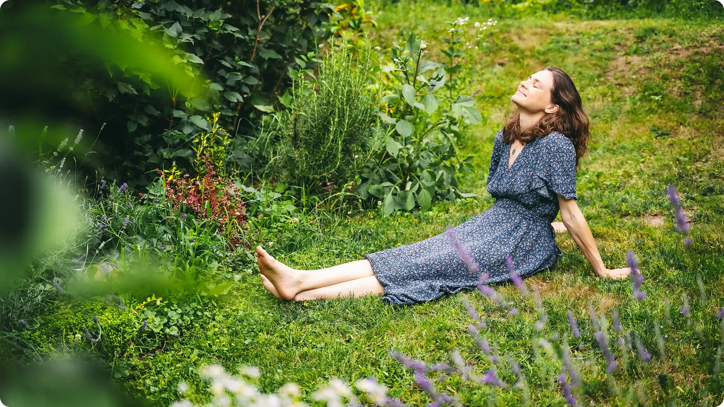 woman in a dress sitting in the grass in a field with her head lifted up to the sky closing her eyes enjoying the space around her