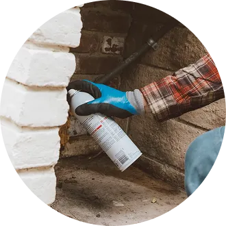 A gloved hand spraying pest control aerosol foam around a foundation crack near brickwork to seal pests out.