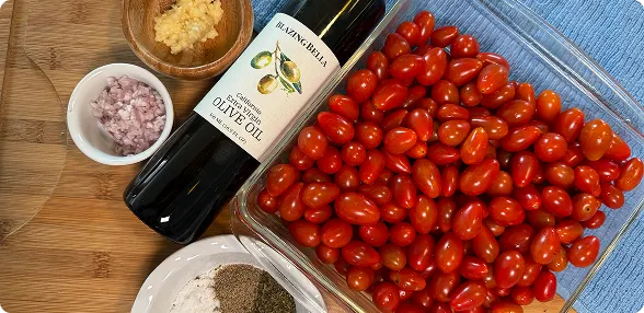 op-down photo of fresh red cherry tomatoes spread on a surface next to a bottle of olive oil and small bowls of seasonings, alongside an editorial article about a viral tomato recipe by Tim Clowers. 