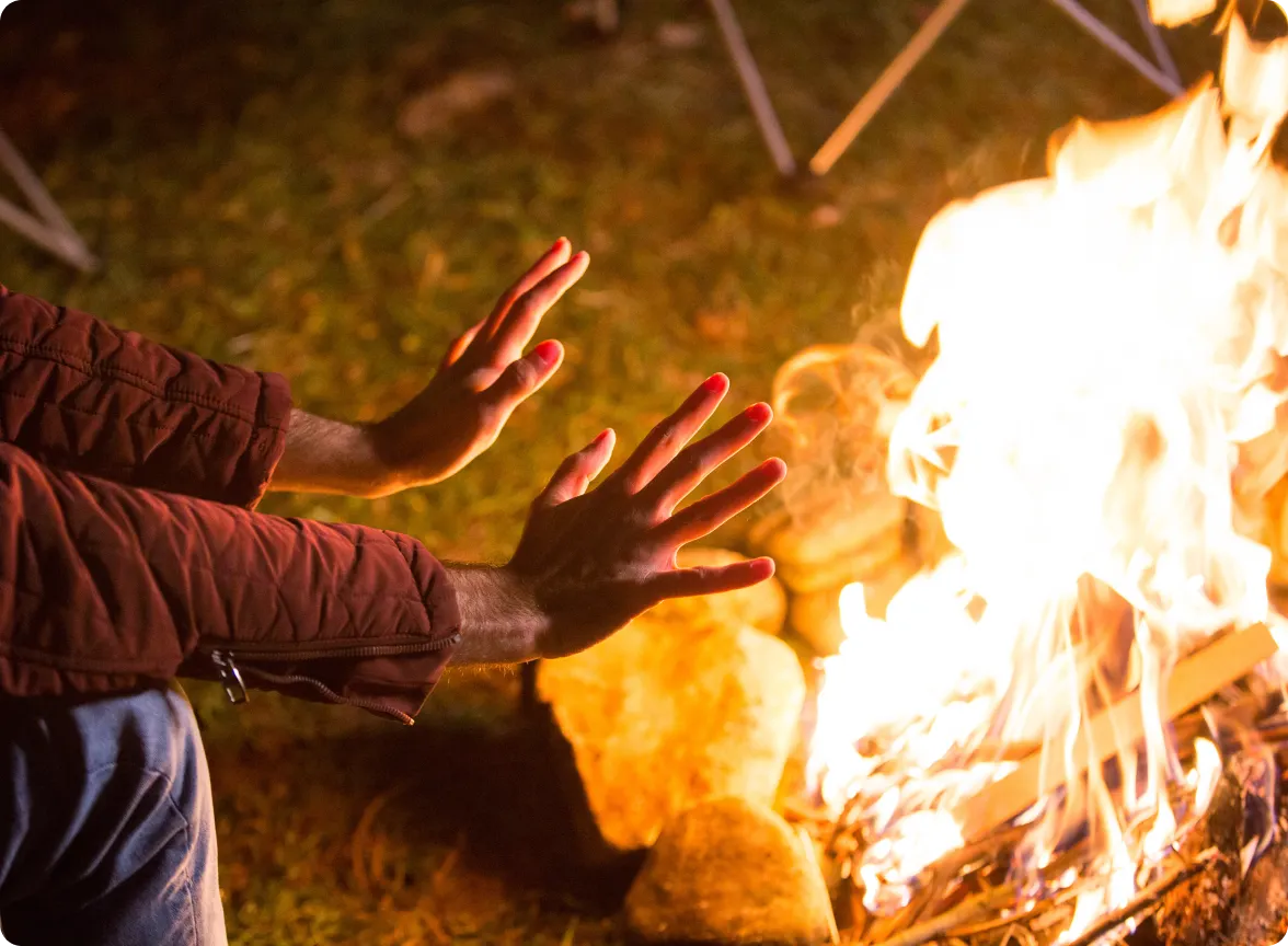 Person warming their outstretched hands near the glowing heat of a burning outdoor campfire.