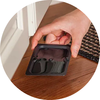 A hand placing a black plastic pest bait trap on a hardwood floor near a doorway for indoor pest control.