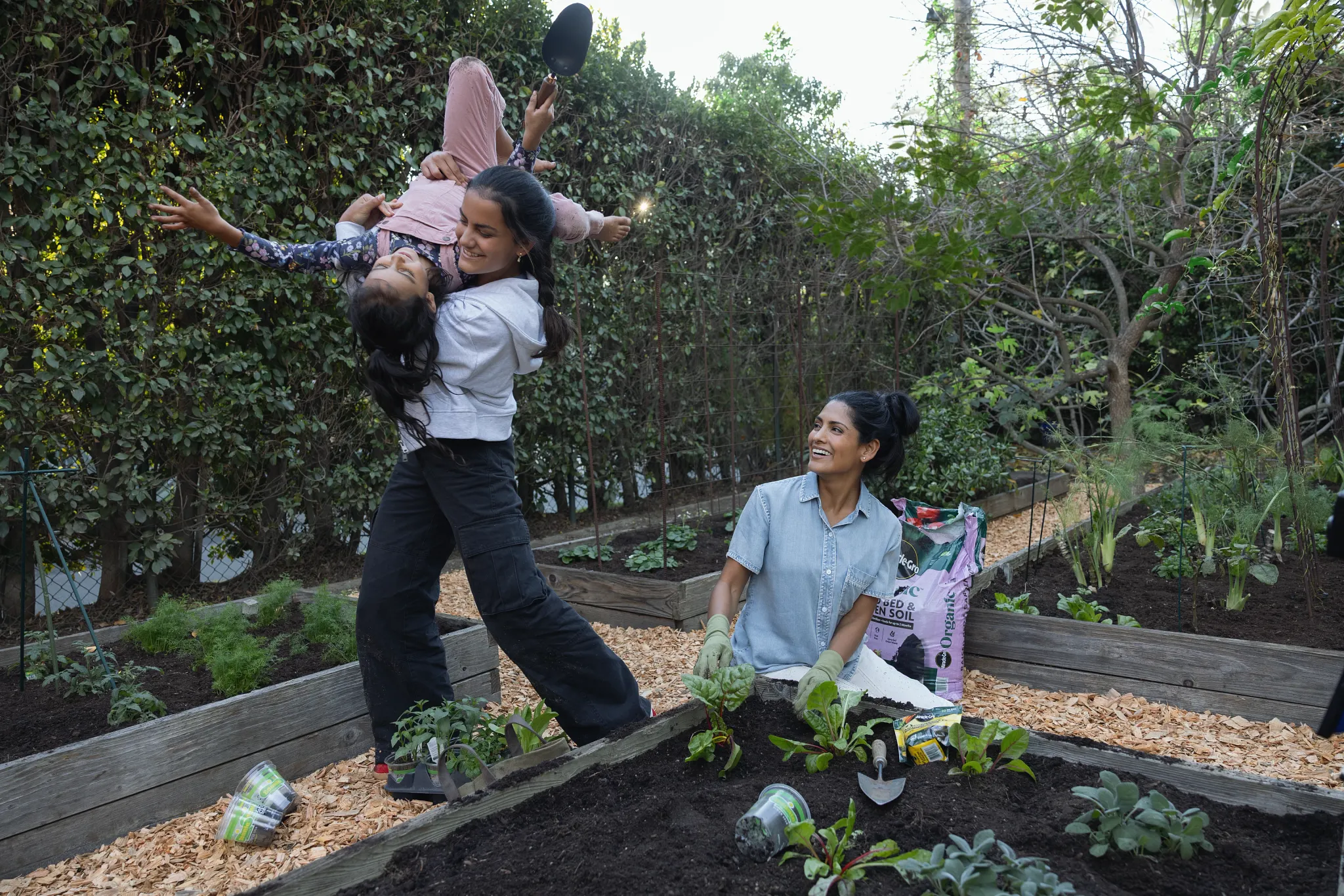 A mother working in a large multi-raised bed garden looks on while her older daughter is picking up and turning her younger sister upside down. The two girls look to have been helping their mom plant new leafy greens. The garden is surrounded by hedge trees and a bag of Miracle-Gro Raised Bed Garden Soil next to their planting position.
