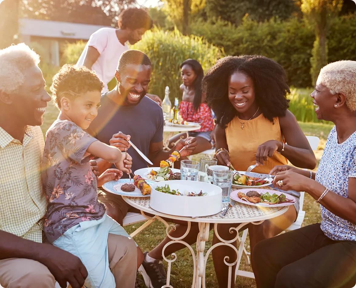 A diverse, multi-generational family sitting and laughing together around an outdoor patio dining table filled with food.