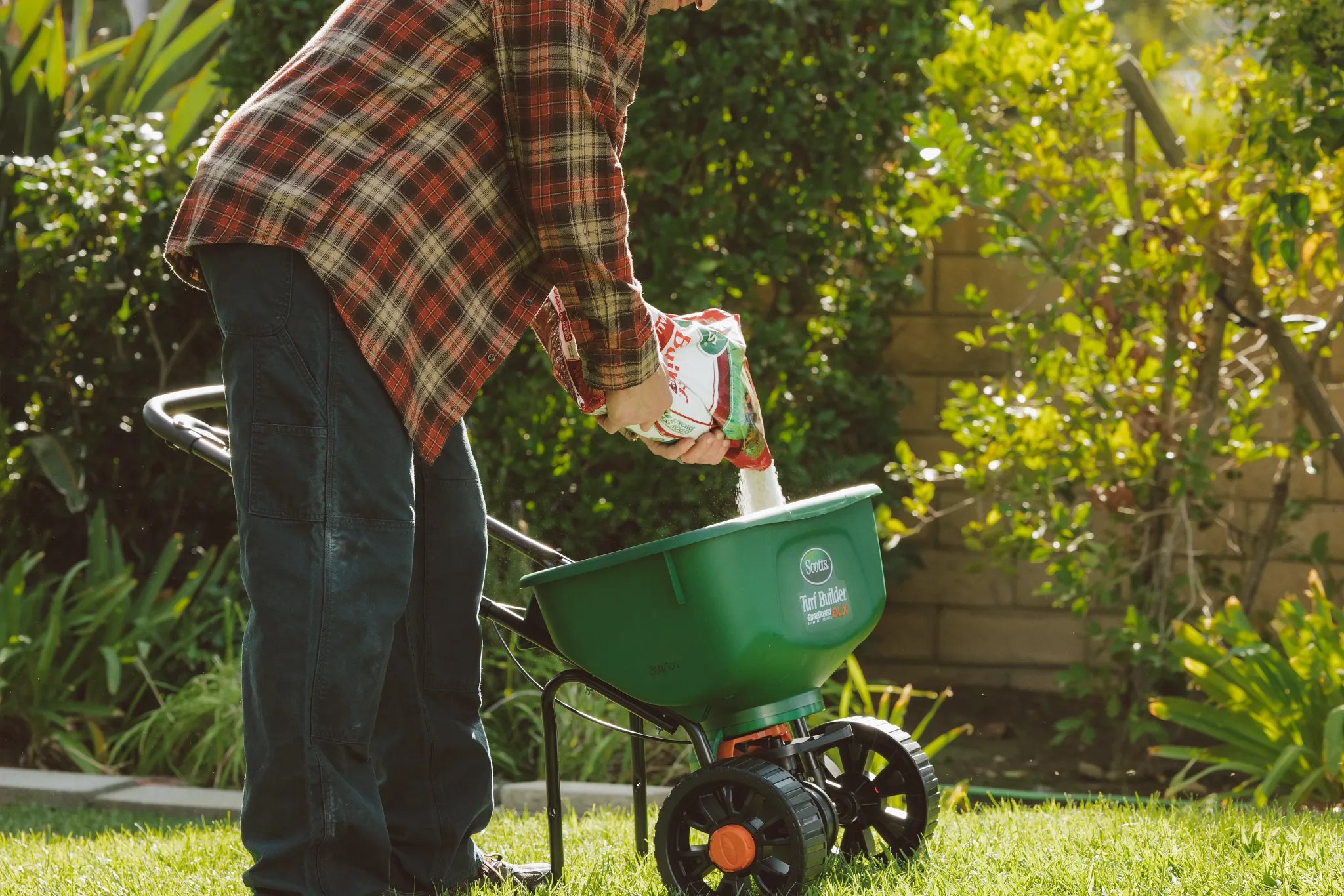 A person in a plaid shirt pours lawn care product from a bag into a green broadcast spreader