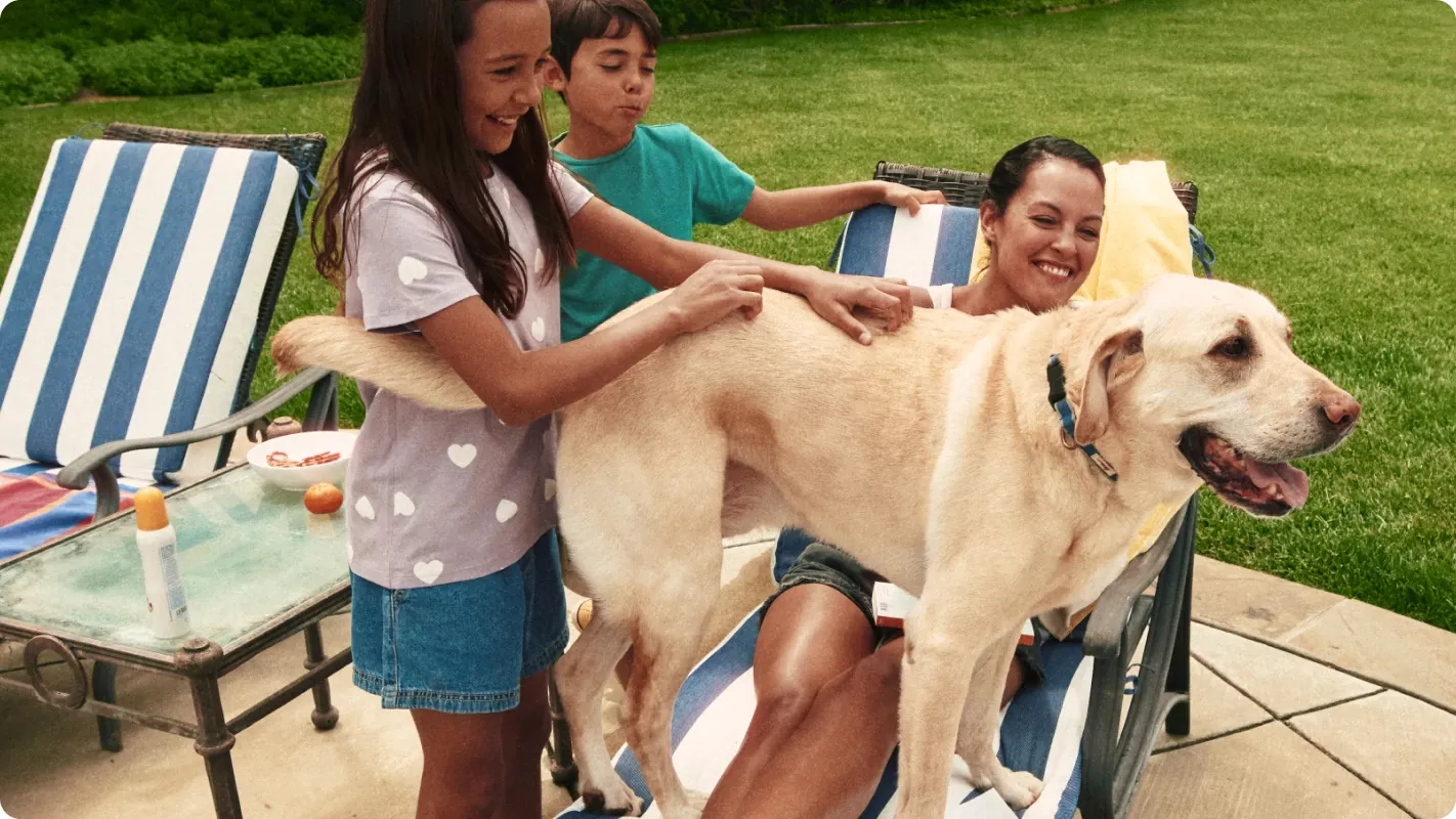 A smiling family petting their yellow Labrador retriever outdoors on a patio next to a lush green lawn.