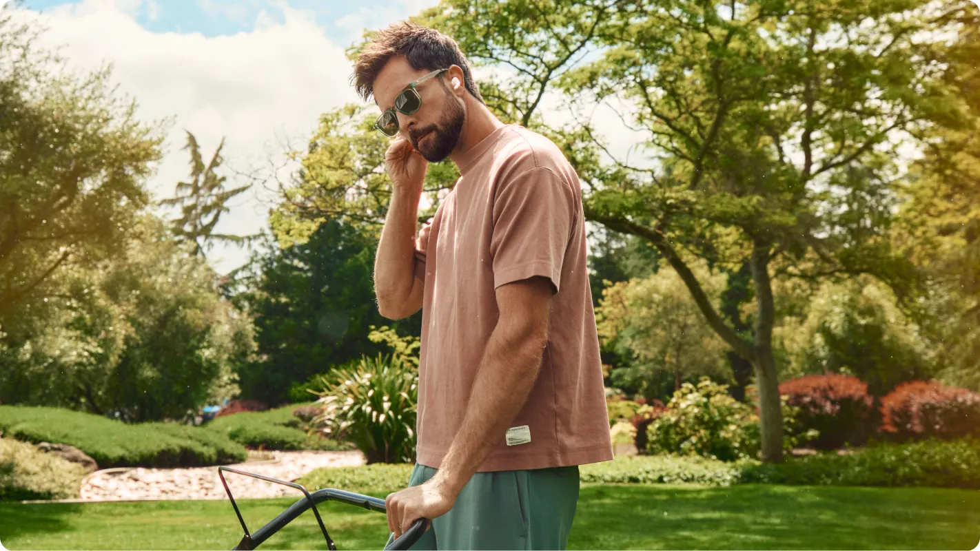 A guy in sunglasses in a t-shirt and shorts reaches up to put an ear bud in his right ear while pushing his lawn mower, all to the backdrop of a beautiful yard with lush green grass multiple trees and bushes surrounding the lawn.