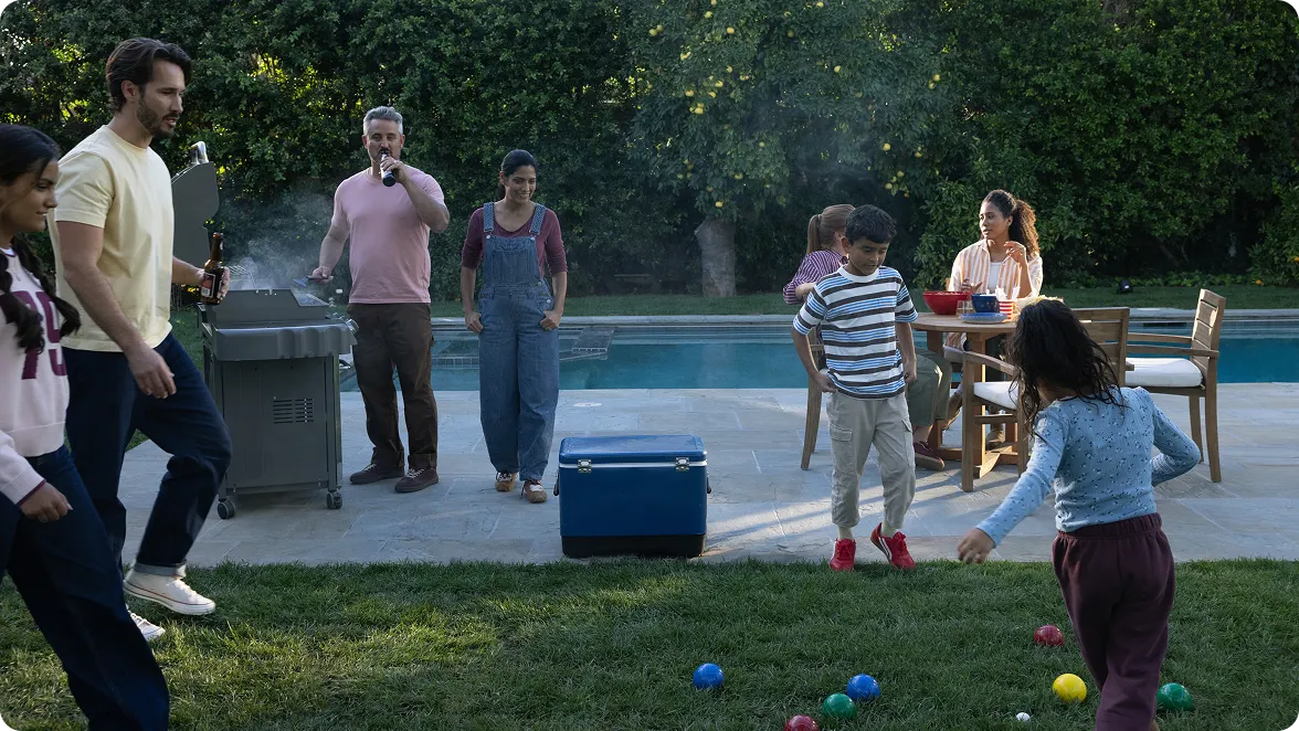 A wide outdoor shot of a multi-generational family enjoying a backyard gathering. In the foreground, children play a game of bocce on a green lawn. In the background, adults stand near a smoking grill and sit at a wooden patio table by a swimming pool. A blue cooler sits on the stone patio, and lush green trees surround the yard. The atmosphere is casual and sunny.
