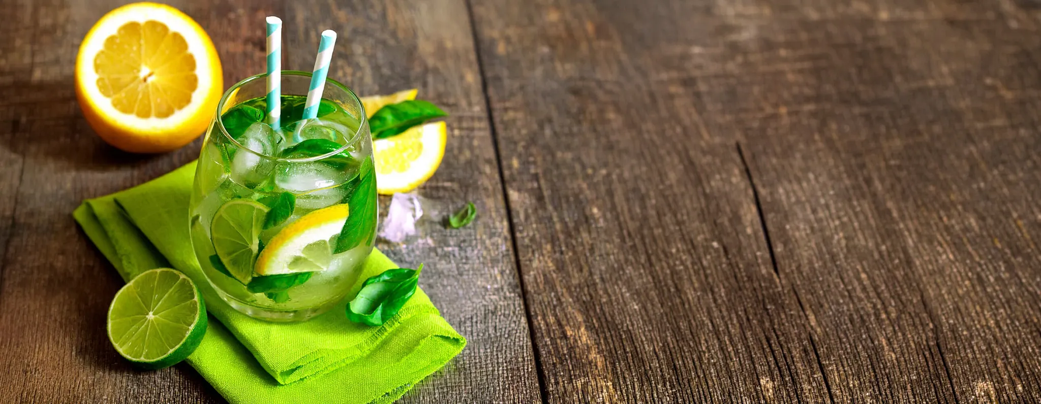 A drink with lemon, lime and mint in a stemless wine glass sits on a wooden table on top of a green napkin with fresh lemon and lime slices next to it.