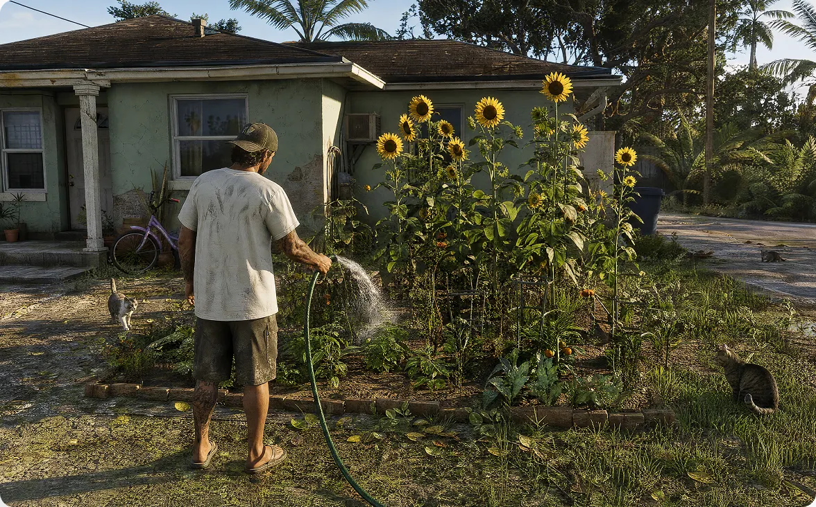 A person standing in a front yard next to bright yellow blooming sunflowers, a small house, and green lawn.