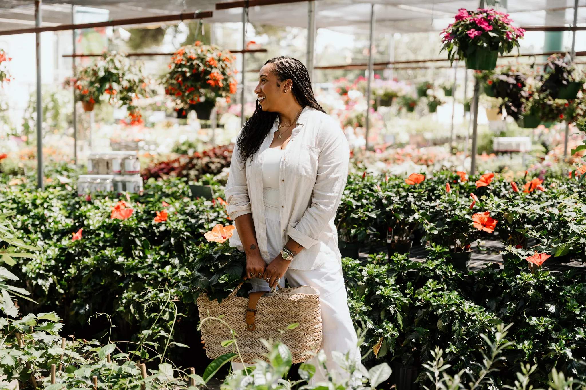 Britt is looking off to the side dressed in a white button down shirt and white slacks standing in the middle of a plant store surrounded by multiple flowering plants and hanging baskets