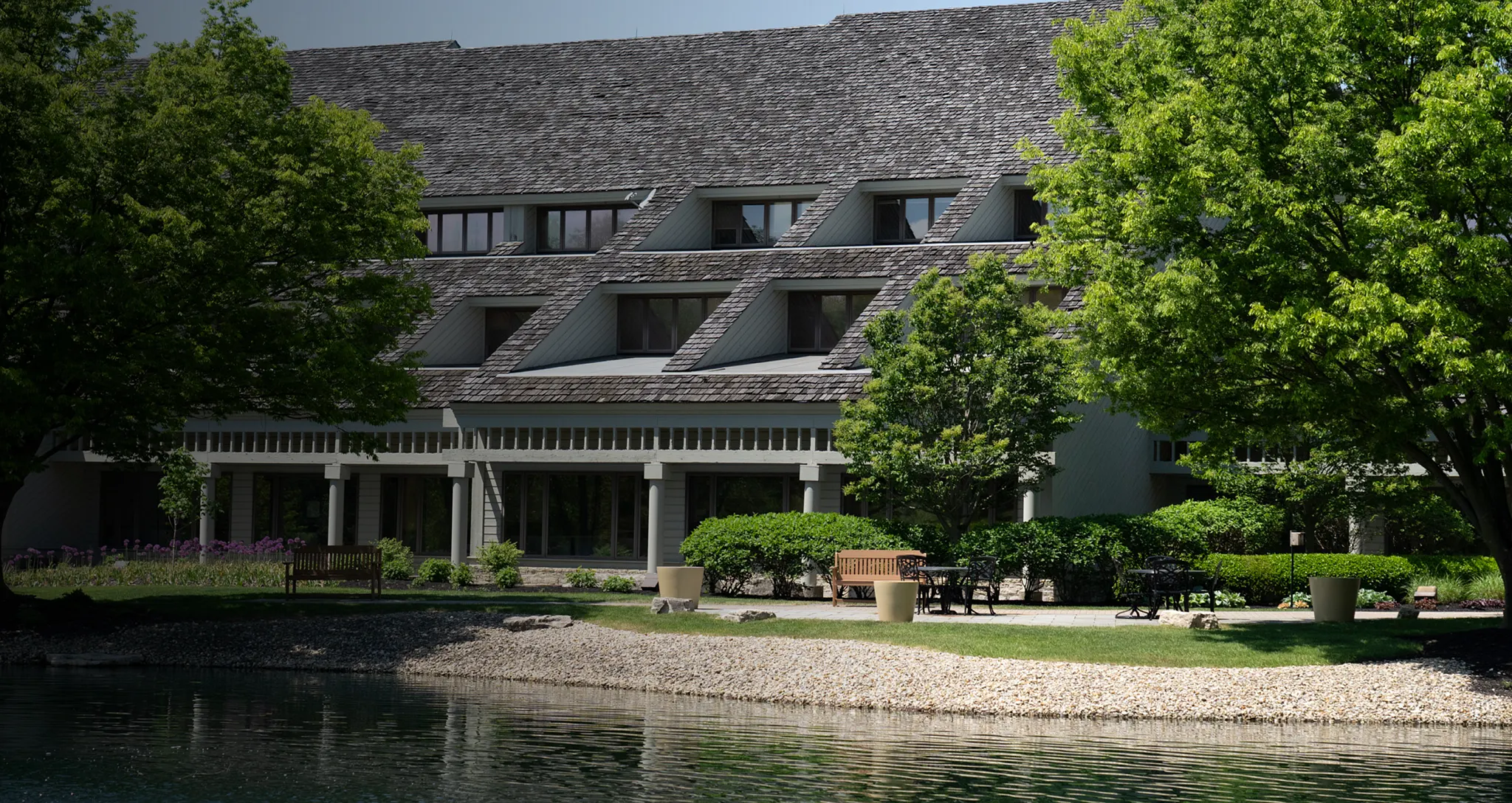 Exterior view of a large, multi-story lodge featuring a dark wooden shingle roof with angled dormer windows. In front of the building is a paved patio area with outdoor seating, potted plants, and manicured green bushes and trees, overlooking a calm body of water with a gravel shoreline in the foreground.