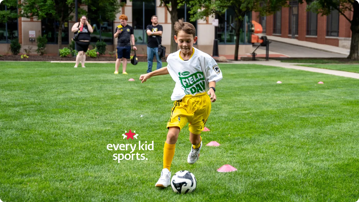 A young boy running and dribbling a soccer ball through small pink cones on a green grass field. He wears a white jersey, yellow shorts, and yellow socks. In the foreground, a white "every kid sports." logo with a small red star graphic is overlaid on the grass.