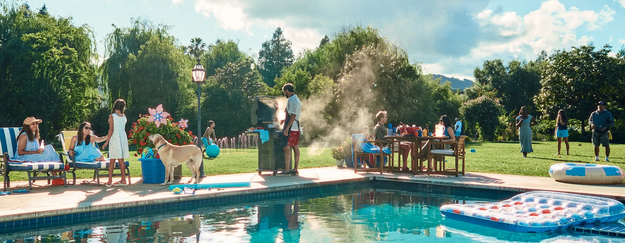 A backyard scene of multiple families hanging out by the pool, grilling and in the yard playing games 