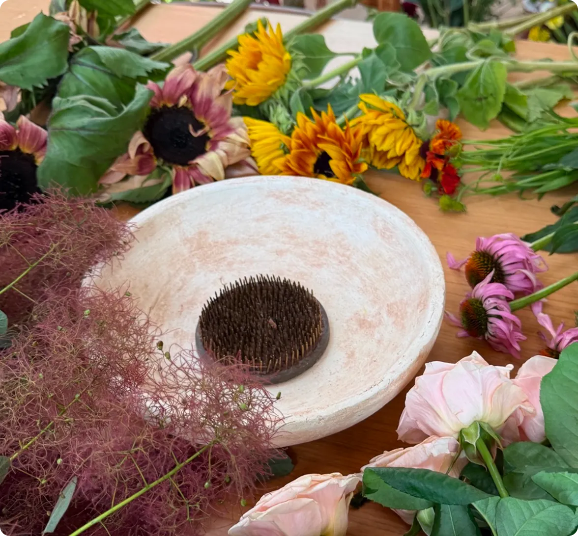 A white, textured shallow bowl containing a metal kenzan flower frog rests on a table surrounded by cut stems, pink coneflowers, and sunflowers, alongside article sections titled 'First, get a flower frog' and 'Next, pick a vase.