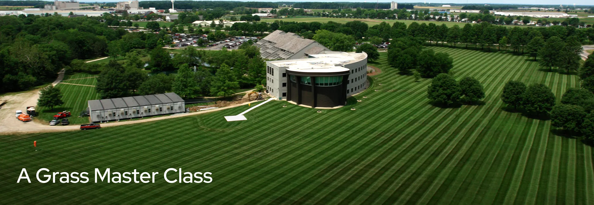 Aerial view of the ScottsMiracle-Gro campus showing a modern building surrounded by a vast, manicured lawn with distinct mowed stripes, trees, and an outer dirt path. Overlay text reads: "A Grass Master Class."