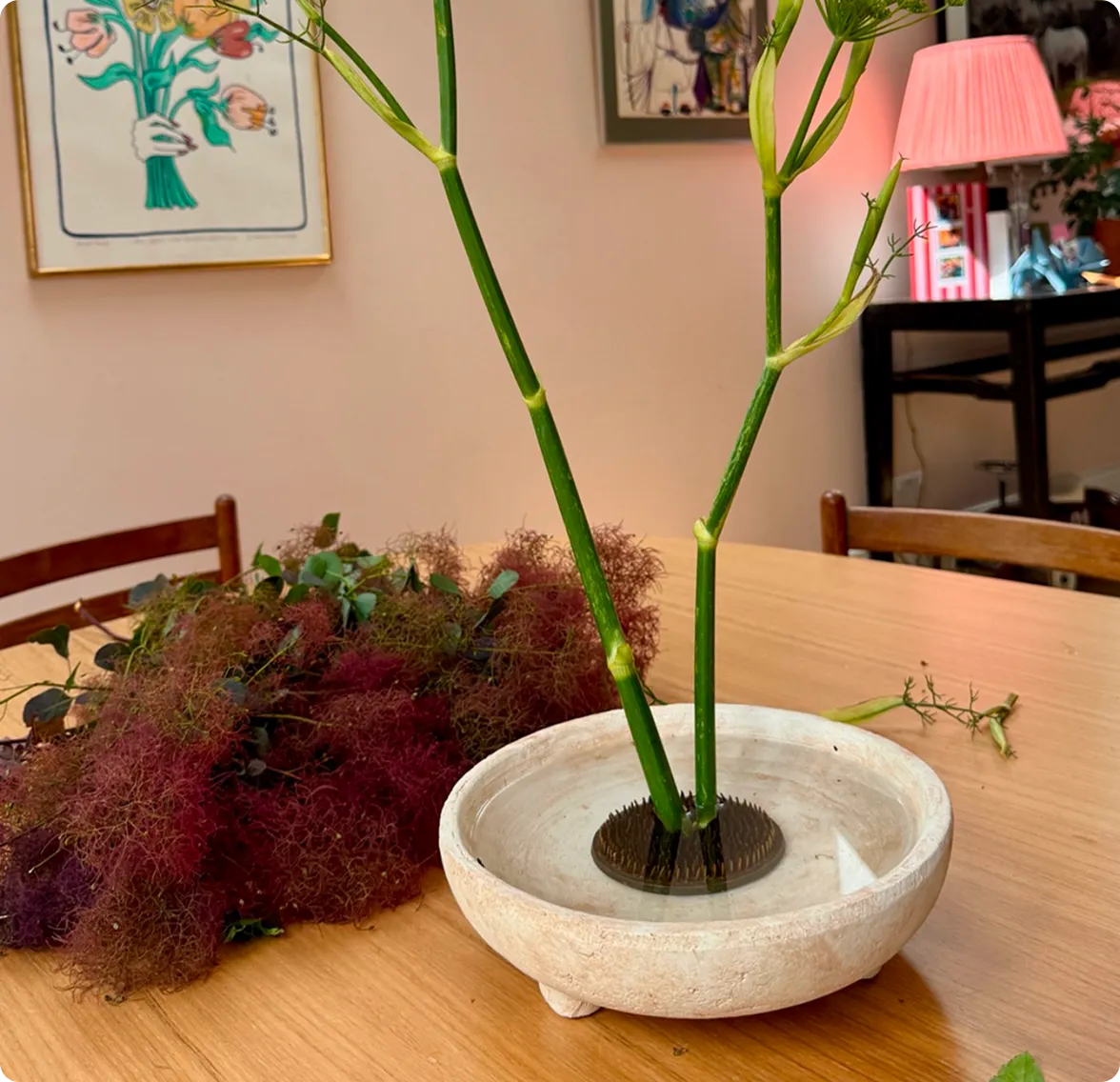 A tall green stem anchored in a water-filled ceramic kenzan bowl on a wooden table, displayed alongside article text titled 'Time to arrange' and 'A word about color.