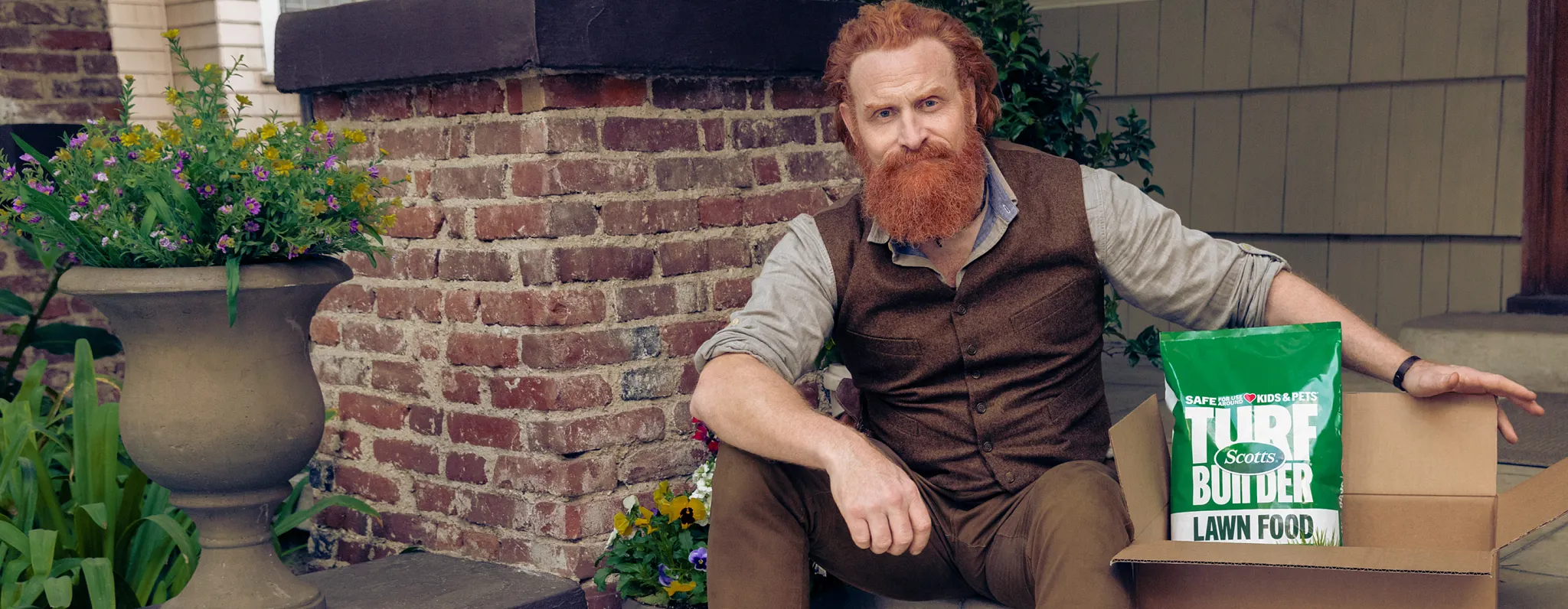 Scottie sitting on the front steps of a brick house with a box open next to him with a bag of Turf Builder Lawn Food showing . 