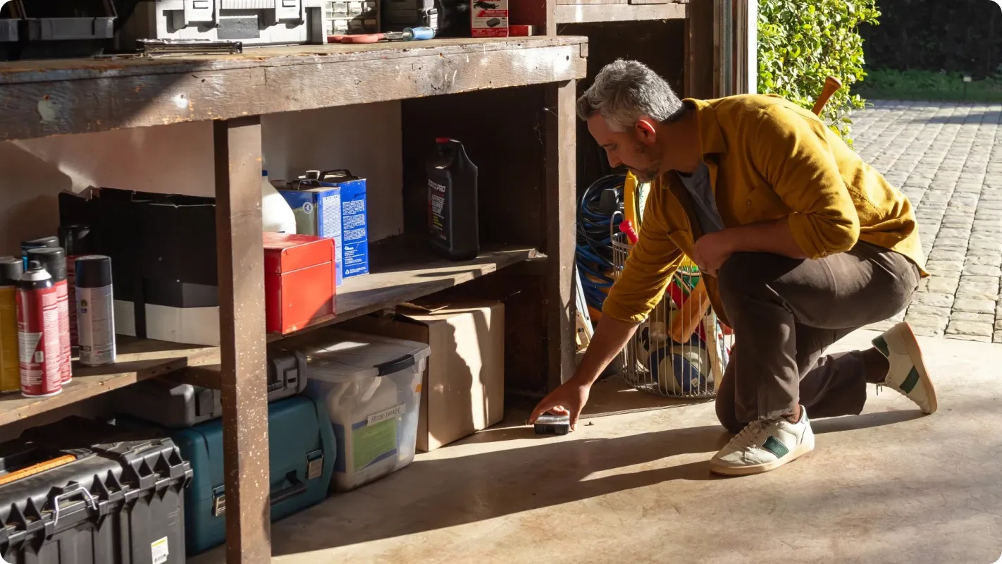 Man placing a rodent bait station under a garage workbench near outdoor entryways.