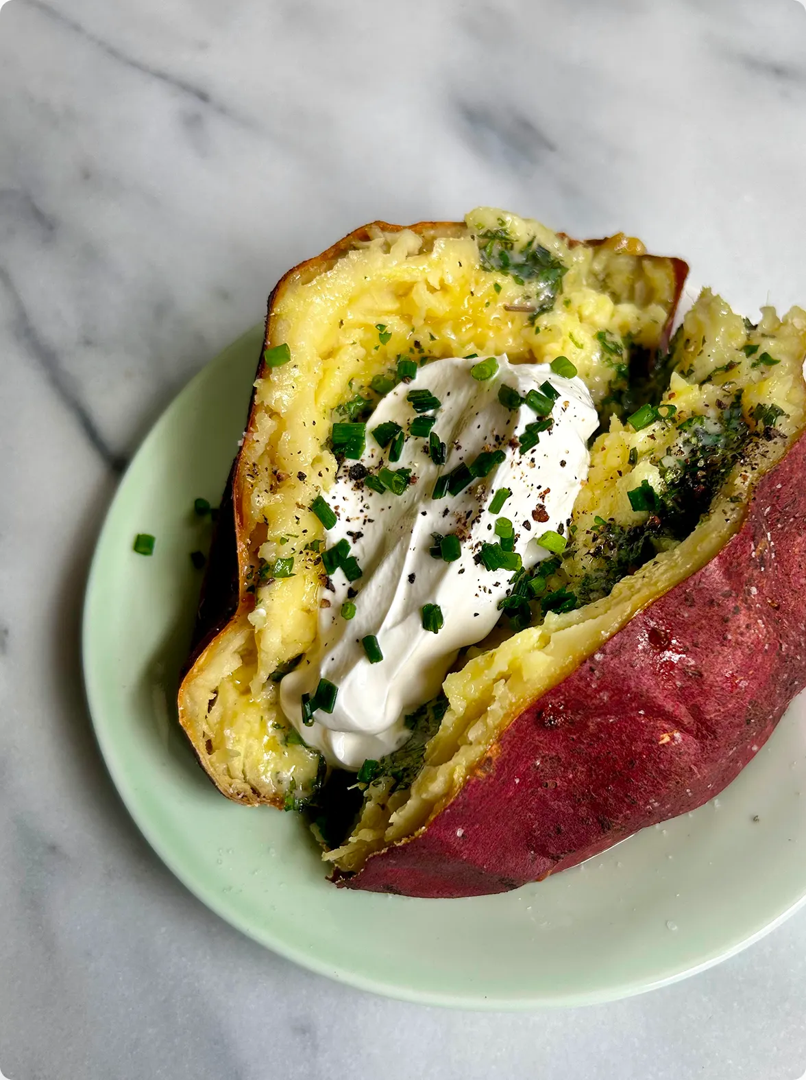 A recipe card titled "Recipe: Baked japanese sweet potatoes with herb butter" features detailed preparation instructions and ingredients on the left. To the right, a top-down view shows a split-open, baked Japanese sweet potato served on a light green plate, topped with green herb butter, a generous dollop of white sour cream, freshly chopped chives, and ground black pepper over a marble background.