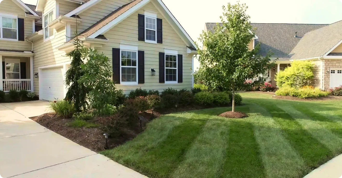 A two-story pale yellow house with black shutters, a concrete driveway, and a neatly manicured green lawn featuring professional mowing stripes.
