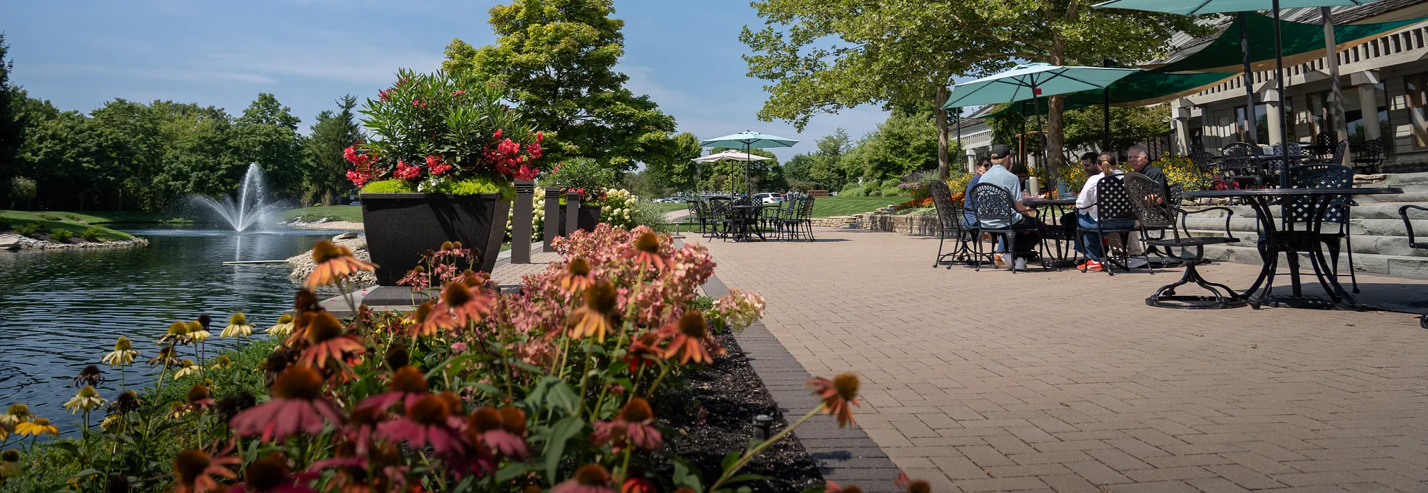 A group of people sitting at outdoor patio tables under umbrellas on the ScottsMiracle-Gro campus, next to a lawn, trees, and pond with a fountain. Overlay text reads: "The man who keeps the ScottsMiracle-Gro campus looking so good. Find out here.
