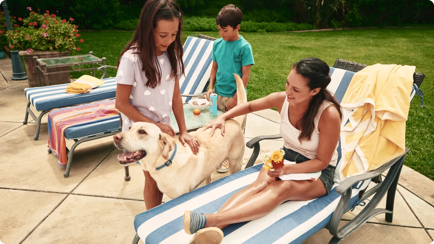 Mom and two kids on the back patio with pool chairs and swim towels laid out are petting their Labrador