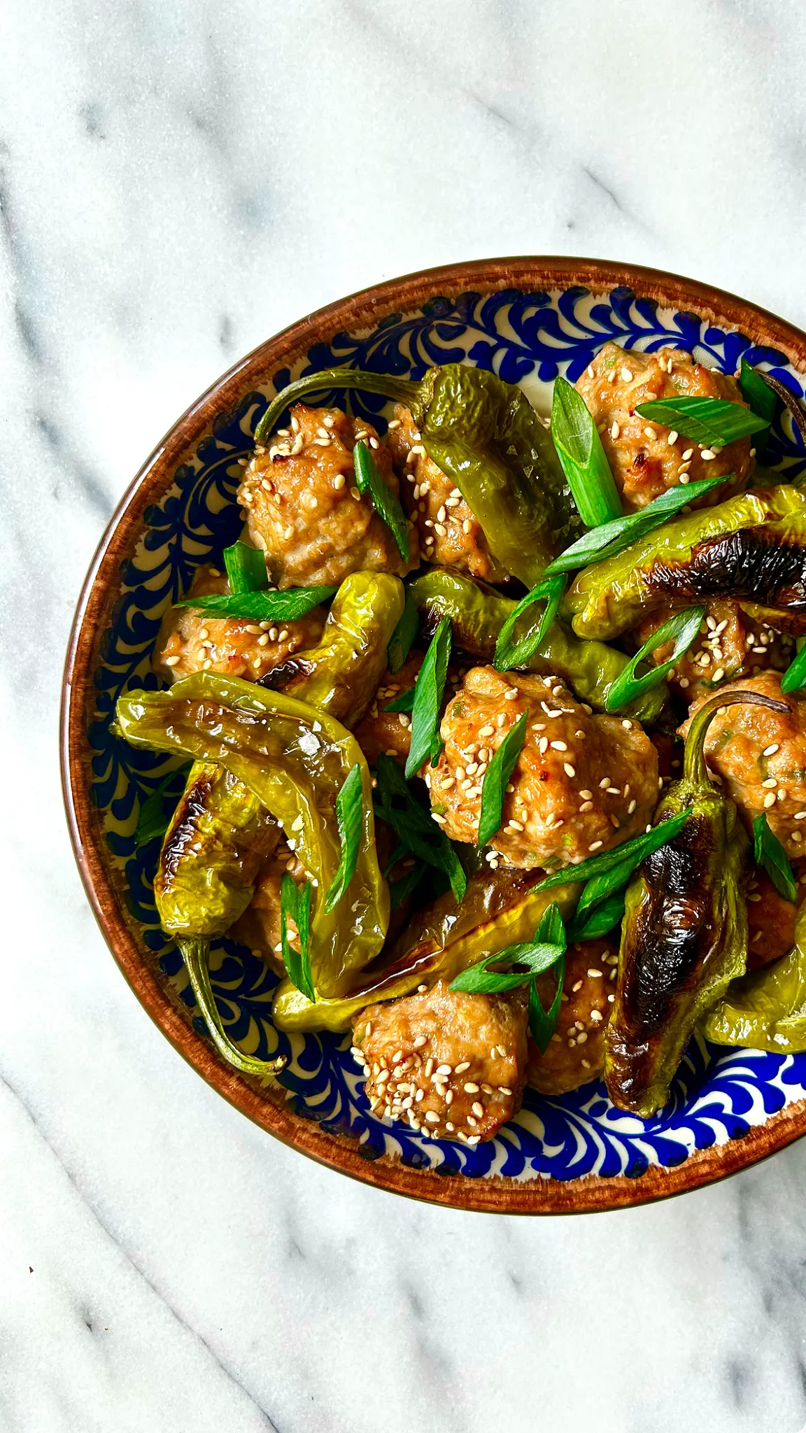A recipe card for Blistered Shishito Peppers and Tsukune-Style Chicken Meatballs features cooking instructions and ingredients on the left. To the right, a top-down view shows a blue-patterned bowl filled with charred green shishito peppers and sesame-seeded chicken meatballs garnished with sliced green scallions on a white marble counter.