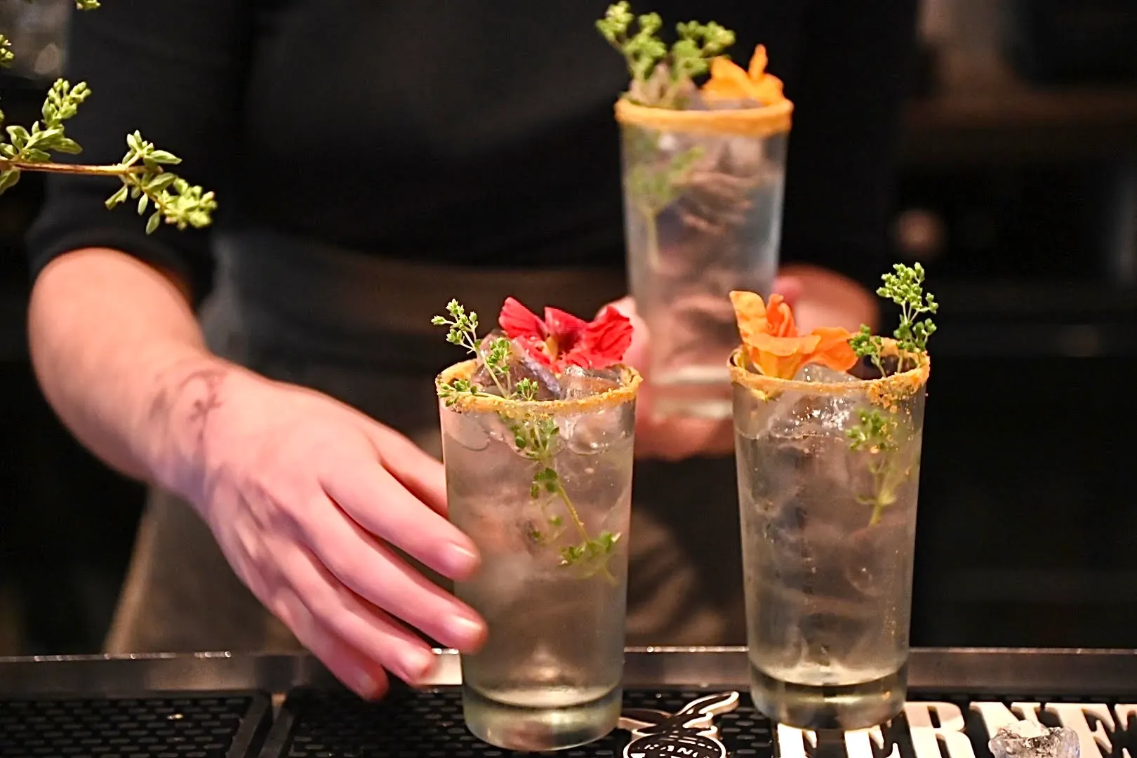 A person&rsquo;s hands serving three clear cocktails in tall glasses on a bar mat. Each drink features a golden-brown rim, ice, a sprig of fresh herbs, and a vibrant edible flower in red or orange. The background is dark and softly blurred, focusing on the colorful floral garnishes.