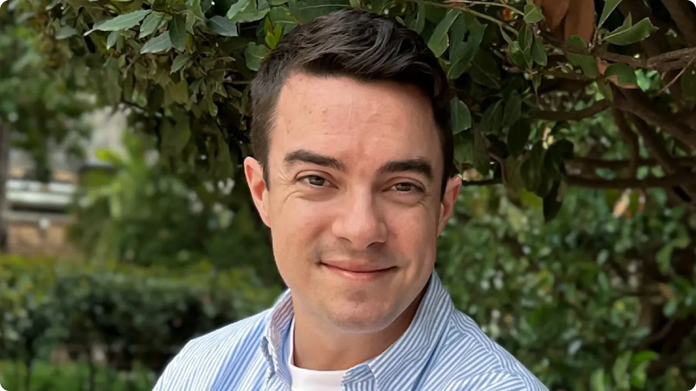 A headshot of Alessandro Pontes smiling outdoors in front of green foliage.