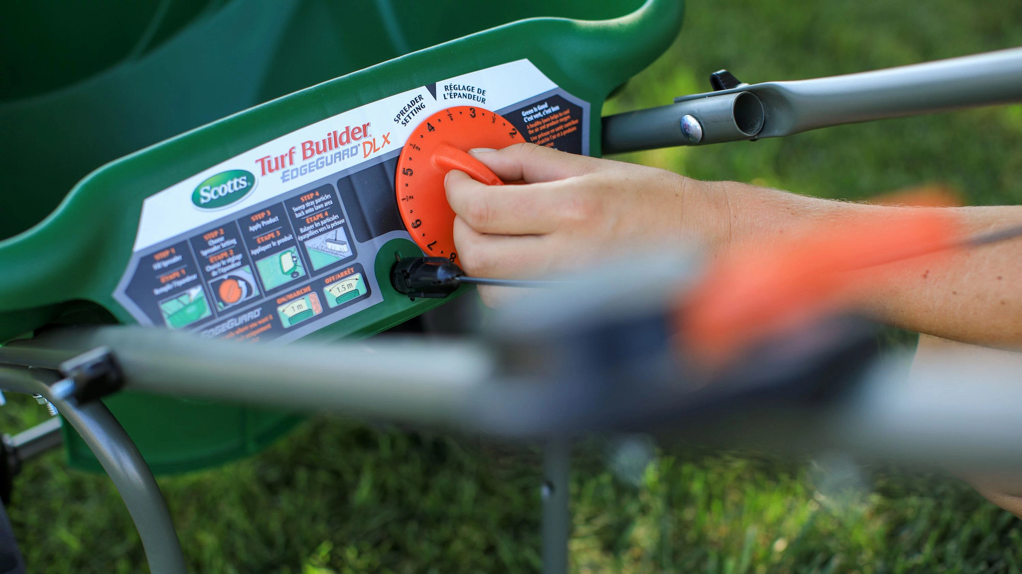 A hand adjusts the orange numbered dial on a green Scotts Turf Builder EdgeGuard DLX lawn spreader.