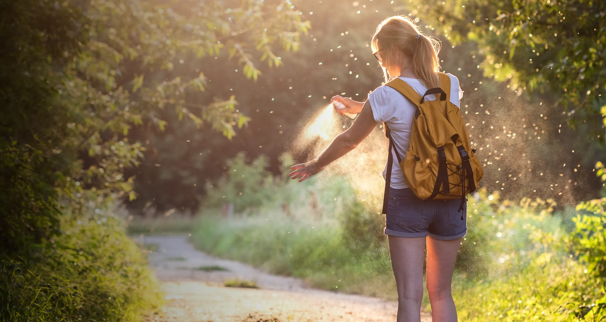 A woman wearing a backpack outdoors, using a hand-held spray bottle to mist her arm against back-lit evening sunlight.
