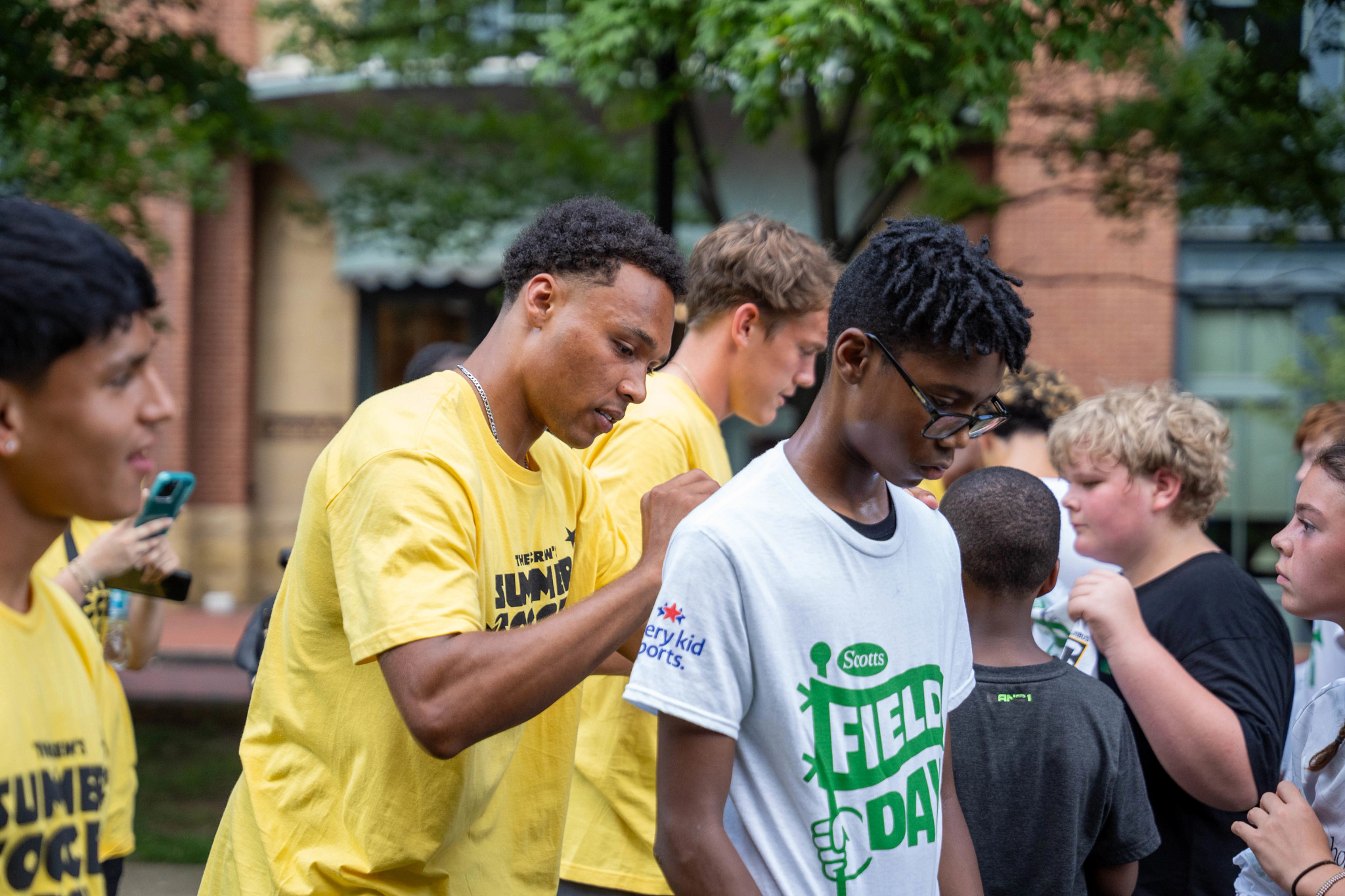 An athlete in a yellow t-shirt signs an autograph on the back of a boy's Scotts Field Day and Every Kid Sports shirt during a youth community event.