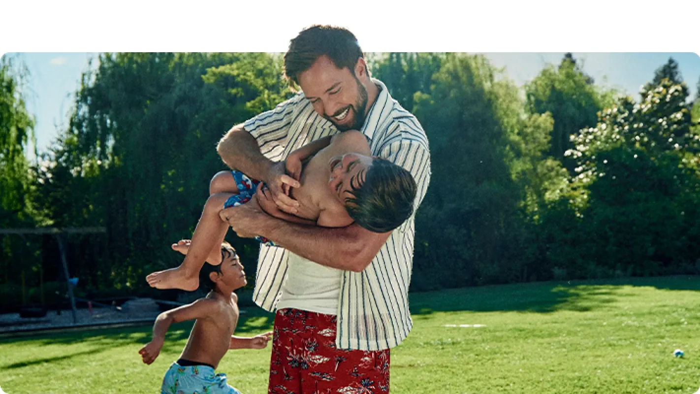 A smiling father playfully holds his young son outdoors in a sunny, grassy backyard.