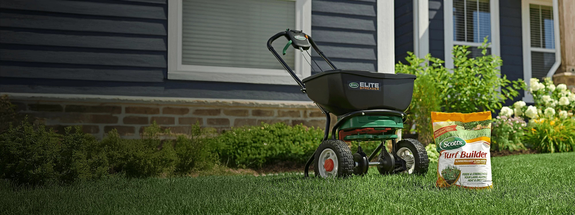 Un &eacute;pandeur Scotts Elite noir est pos&eacute; sur une pelouse verte, &agrave; c&ocirc;t&eacute; d'un sac d'engrais Scotts Turf Builder SummerGuard avec insecticide. &Agrave; l'arri&egrave;re-plan, on aper&ccedil;oit une maison bleu fonc&eacute; avec des finitions blanches.