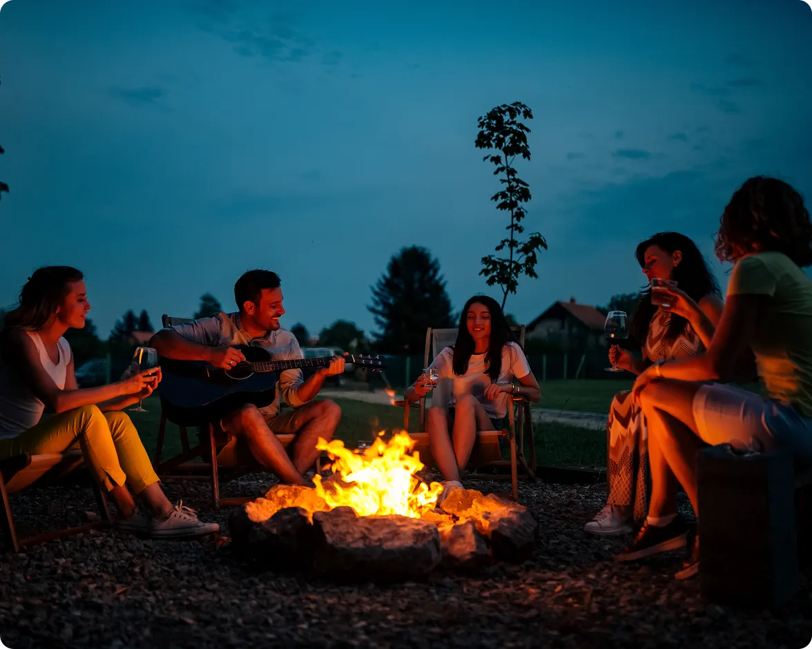 An outdoor night photograph showing a group of people sitting in lawn chairs in a circle around a burning bonfire, with light casting across the group and dark trees visible against a dusk sky in the background.