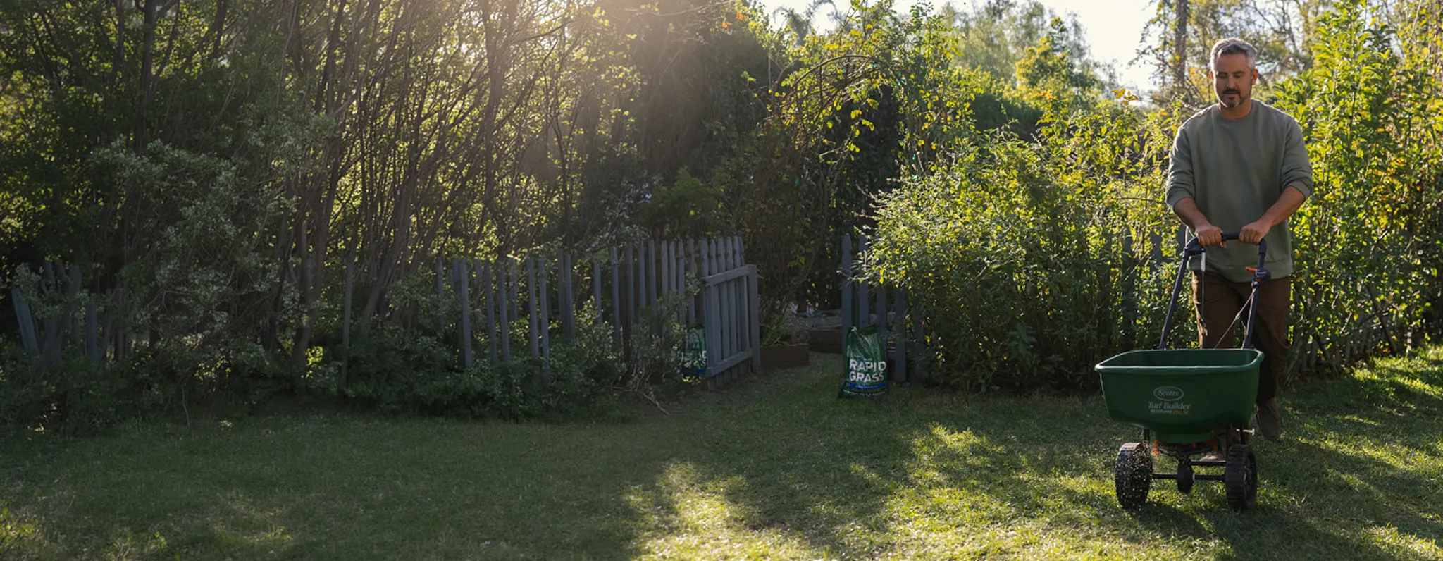 A man using a green lawn spreader in a sunny backyard with lush trees. A large white overlay reads "Limited-Time Deal! Save 20% and get a FREE spreader when you sign up for the annual subscription plan." Includes a black "Save Now" button.