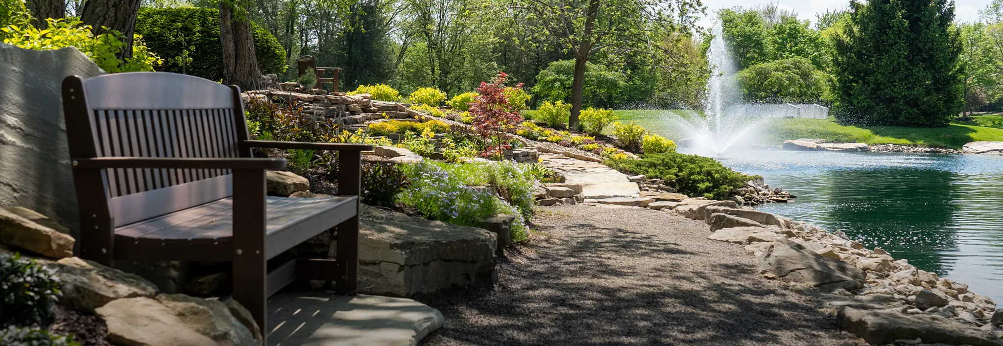 Wooden bench along a gravel walking path beside a landscaped pond with a fountain.