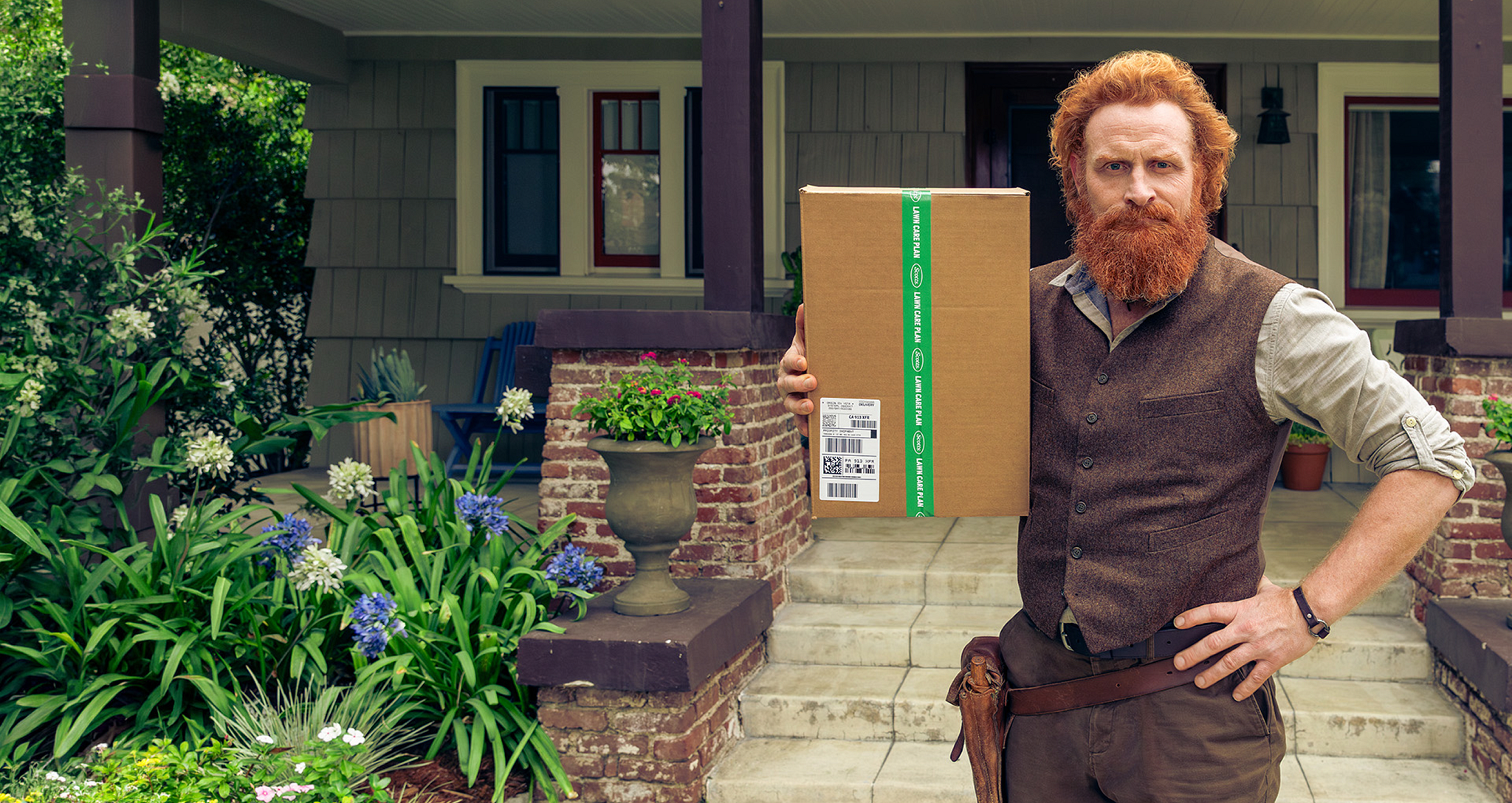 A man with a red beard in a brown vest holding a subscription box for a custom lawn care plan in front of a house with a green garden. Text reads "Take the Guesswork Out of Lawn Care" with a button to "Build Your Lawn Plan.