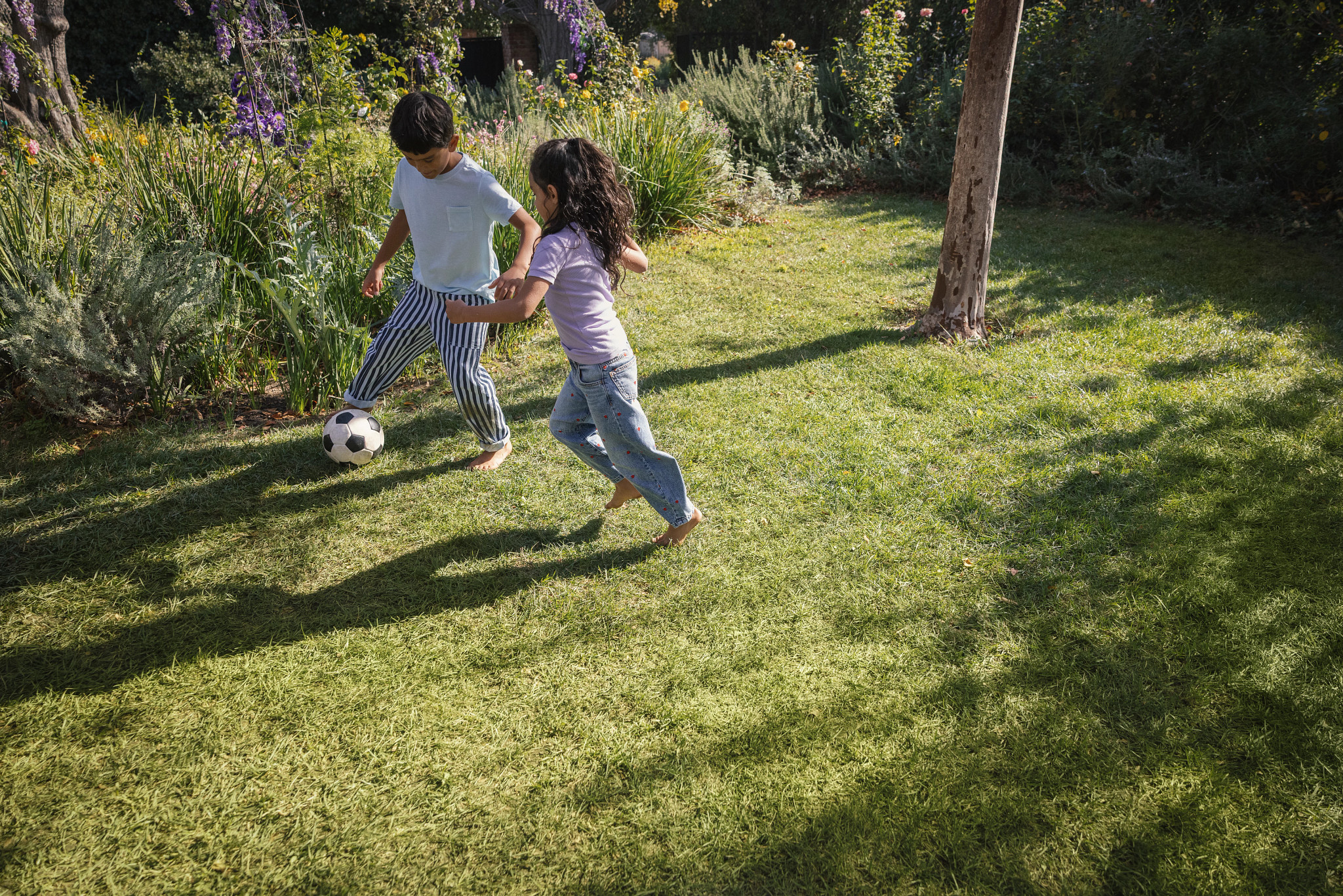 Two young children playing soccer together on a lush green lawn in a sunny backyard garden