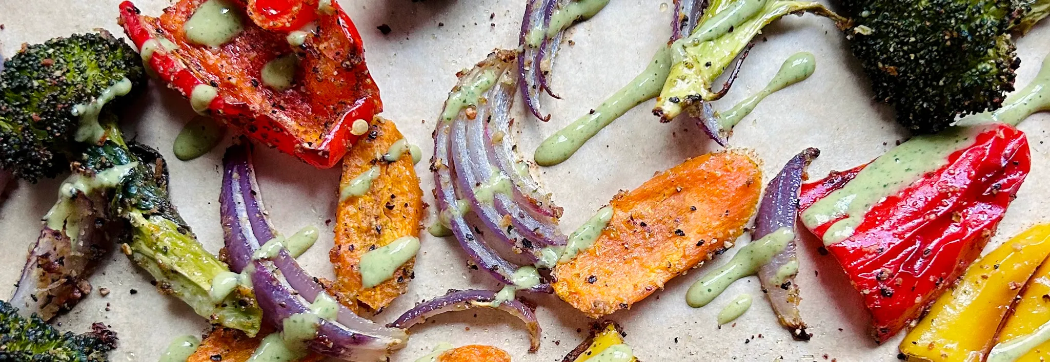 Top-down view of roasted vegetables&mdash;including charred broccoli, red and yellow bell peppers, purple onions, and sweet potatoes&mdash;seasoned and drizzled with a creamy green herb sauce on parchment paper.