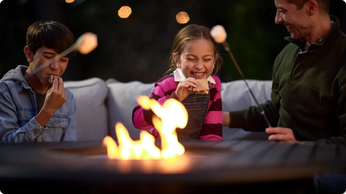 A family roasting marshmallows and making s'mores around an outdoor patio fire pit at night.