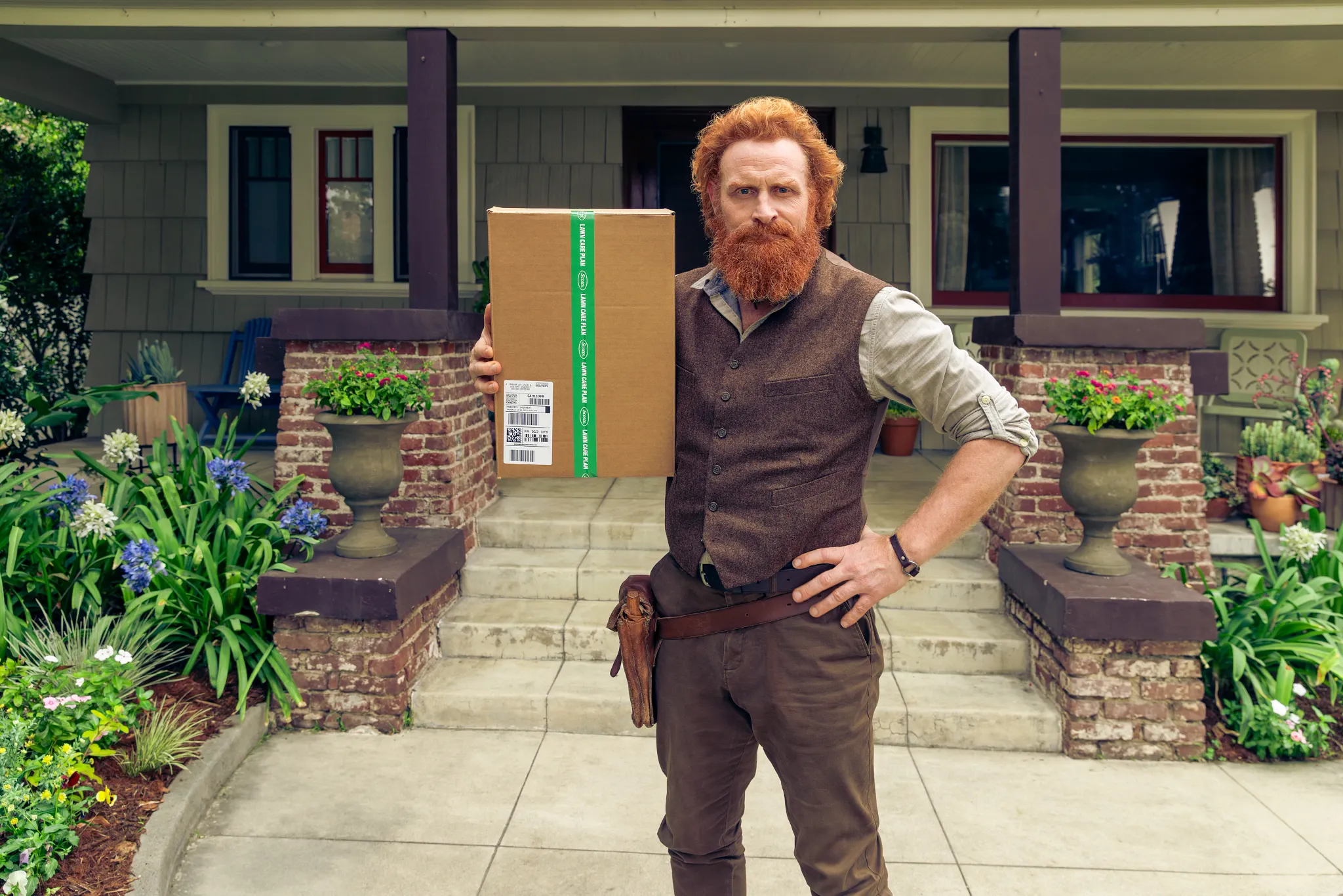 Scott from Scotts standing on a brick walkway holding a cardboard delivery box in one hand.