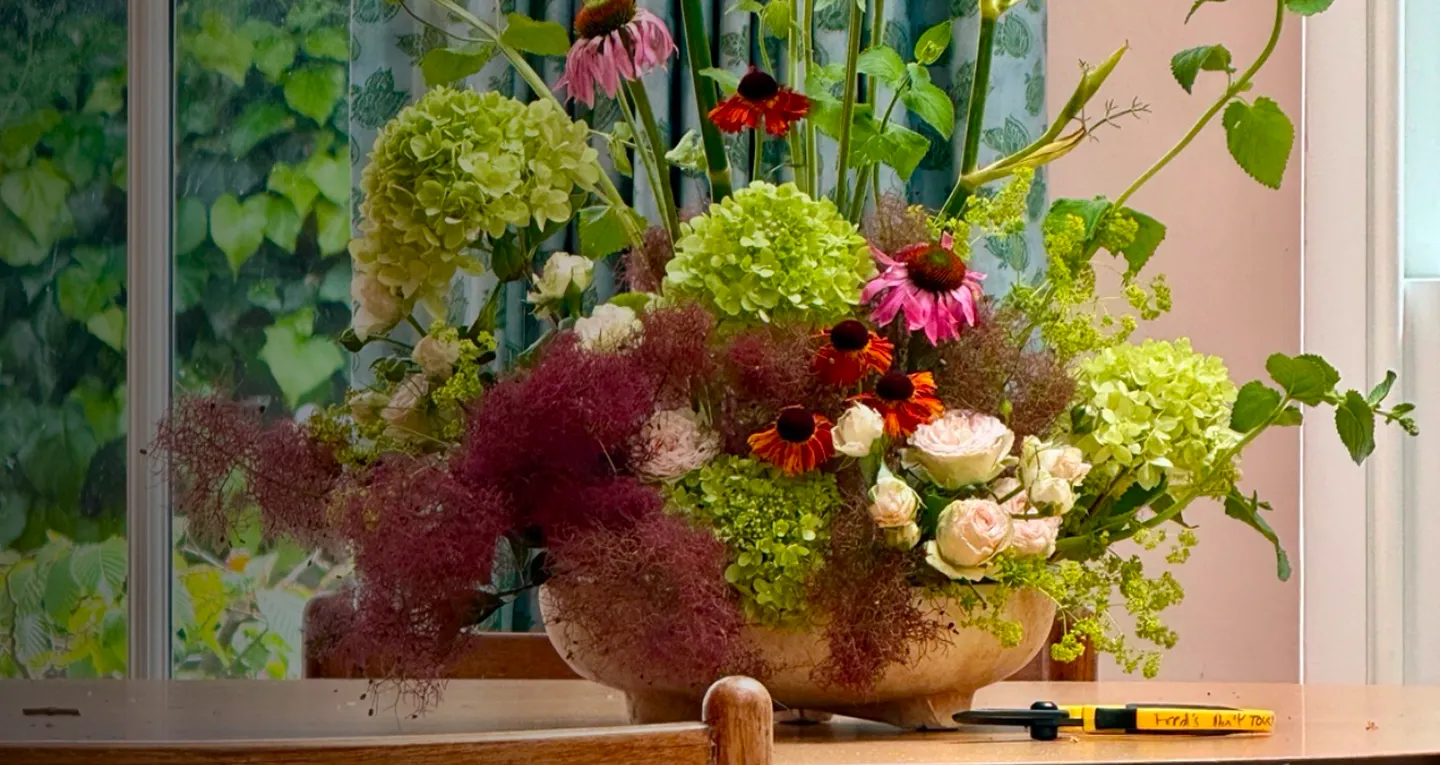A floral arrangement featuring pink coneflowers, green hydrangeas, and dark red smokebush in a shallow ceramic bowl on a wooden table